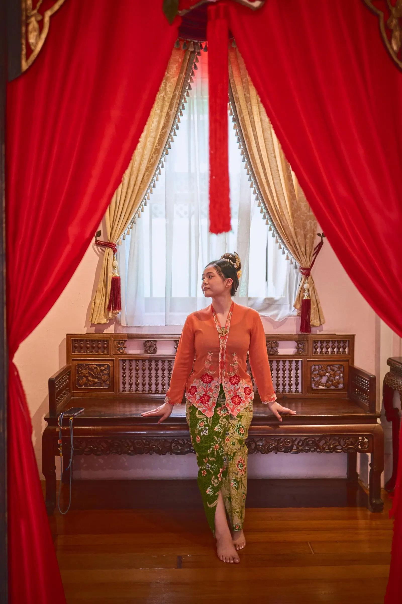 Woman in an orange Nyonya kebaya on a carved bench between red curtains, Penang